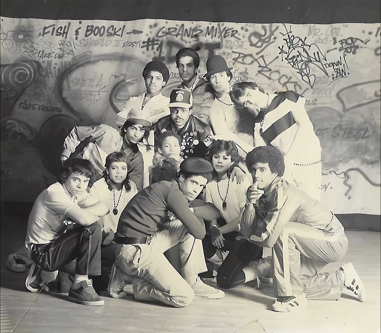 Black and white photo of 12 1980s breakdancers posing in front of a wall with graffiti.