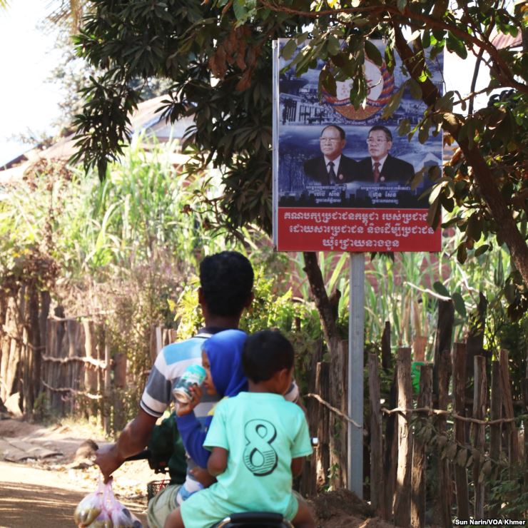 People riding a motorcycle under a campaign sign.