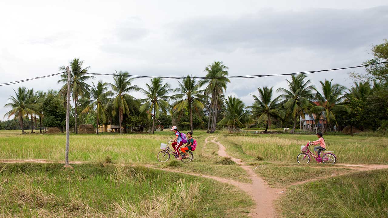 People ride their bikes along a rural dirt road with palm trees along the horizon.