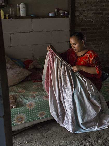 Photo of Rasminah folding a sheet while sitting on a mattress.