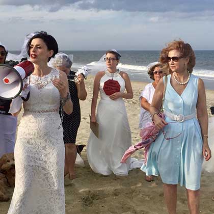 Photo of Fraidy Reiss, left foreground, carrying a megaphone and leading activists wearing wedding gowns across a beach.