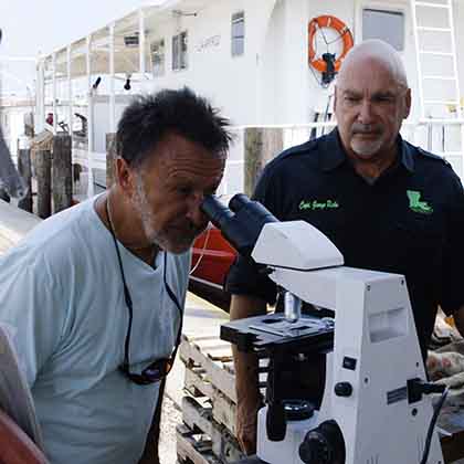 Two men standing on a dock next to a boat. One of them is looking into a microscope. 